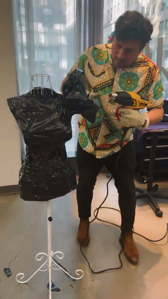 Tony building a gown of black bags on a mannequin, with a heat gun